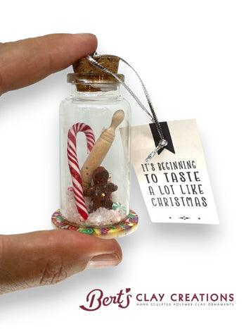 Miniature jar with Christmas-themed items and a tag, held by a hand on a white background. Brand 'Bert's Clay Creations' visible.